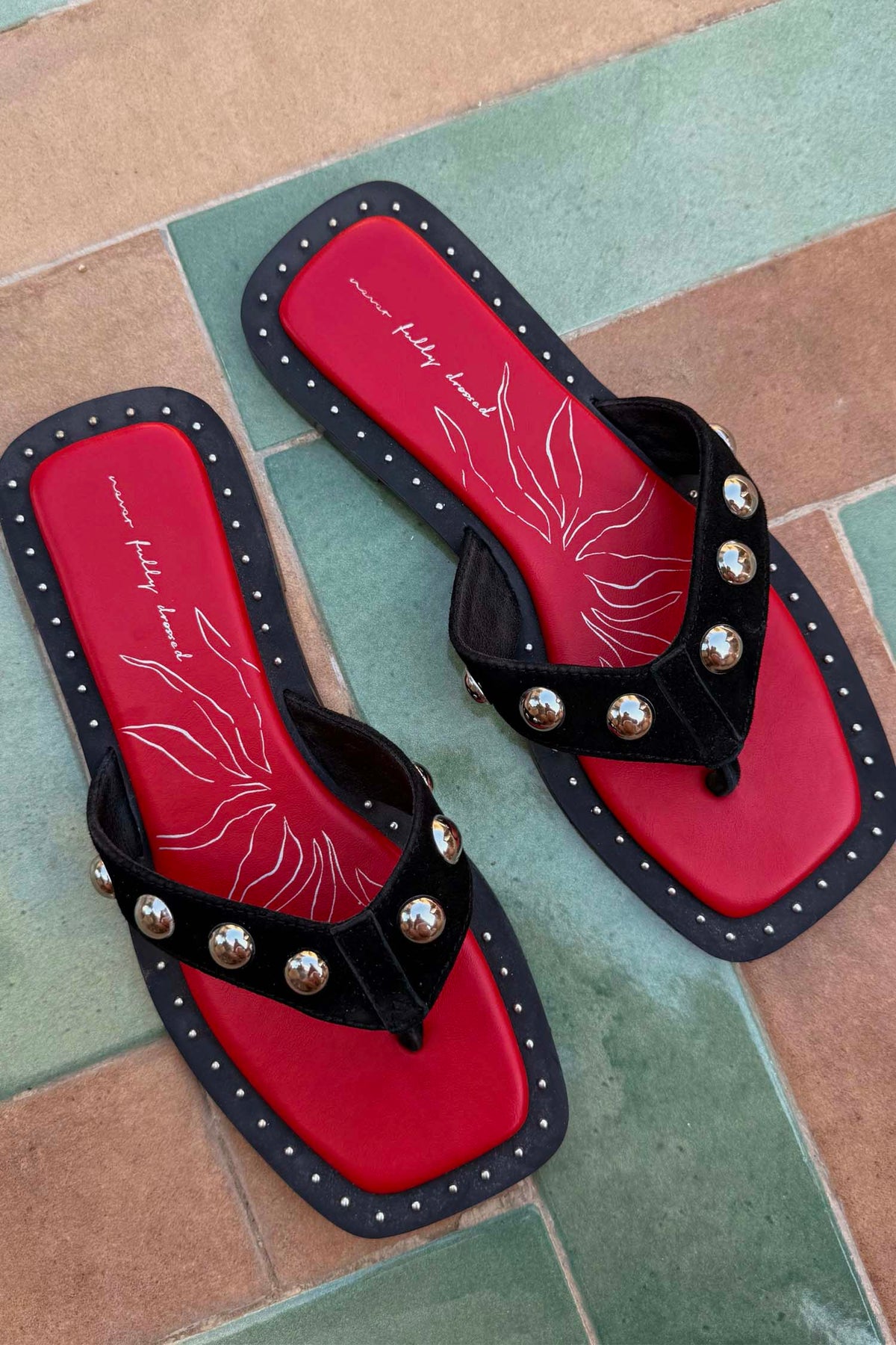 Black suede flat sandals featuring silver studs, a square toe, and a red logo footbed with white graphic details.