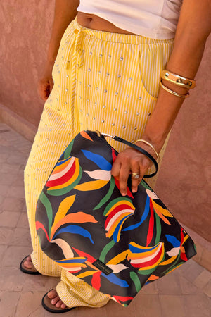 NFD banana print pouch with a black wrist strap, held by a model wearing studded yellow and white striped trousers.