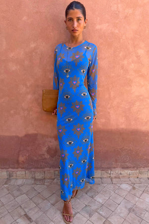 Blue Eye Mesh Maxi Dress with pink heart and eye motifs and long sleeves, worn by a model against a terracotta wall.