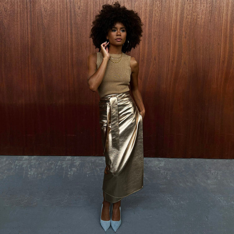 Gold Vegan Leather Jaspre Skirt styled with a sleeveless knit top on a model against a wooden wall.