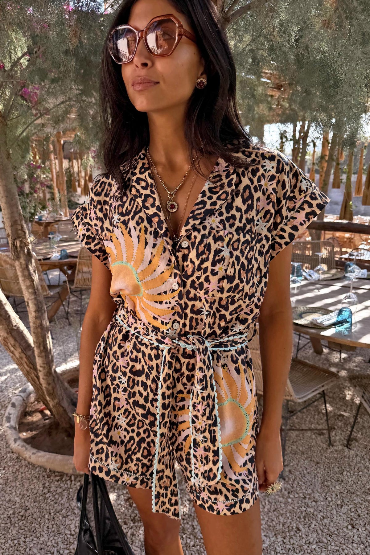 Leopard Print Playsuit with soleil motifs, turquoise trim and a tie waist, worn by a model at an outdoor cafe.