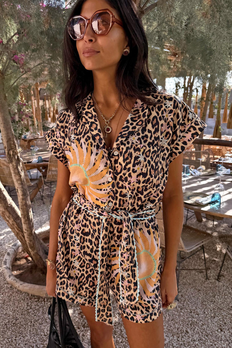 Leopard Print Playsuit with soleil motifs, turquoise trim and a tie waist, worn by a model at an outdoor cafe.