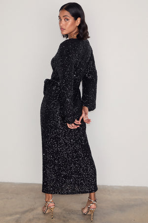 Black Sequin Kira Wrap Dress with long sleeves and a tie waist, shown on a model wearing animal print heels.
