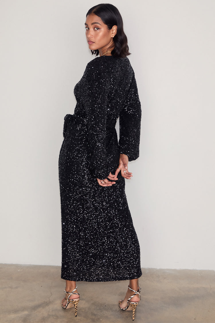 Black Sequin Kira Wrap Dress with long sleeves and a tie waist, shown on a model wearing animal print heels.