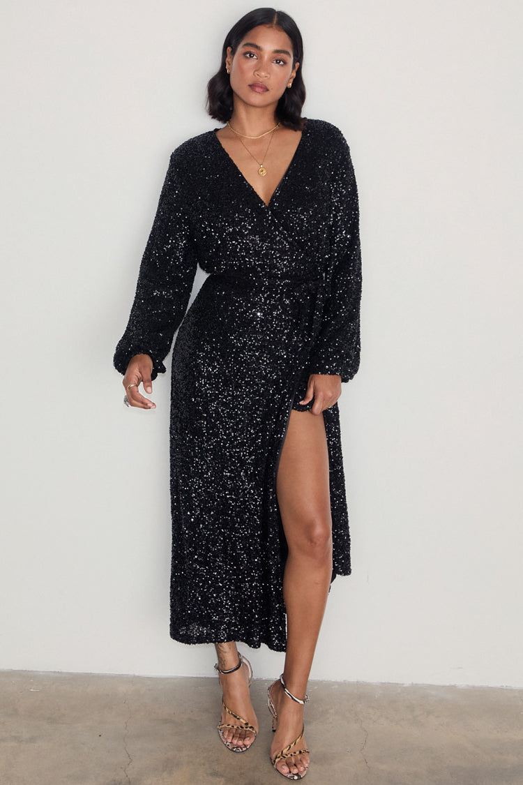 Black Sequin Kira Wrap Dress featuring a multi-wear wrap style, long sleeves, and a leg slit on a model.
