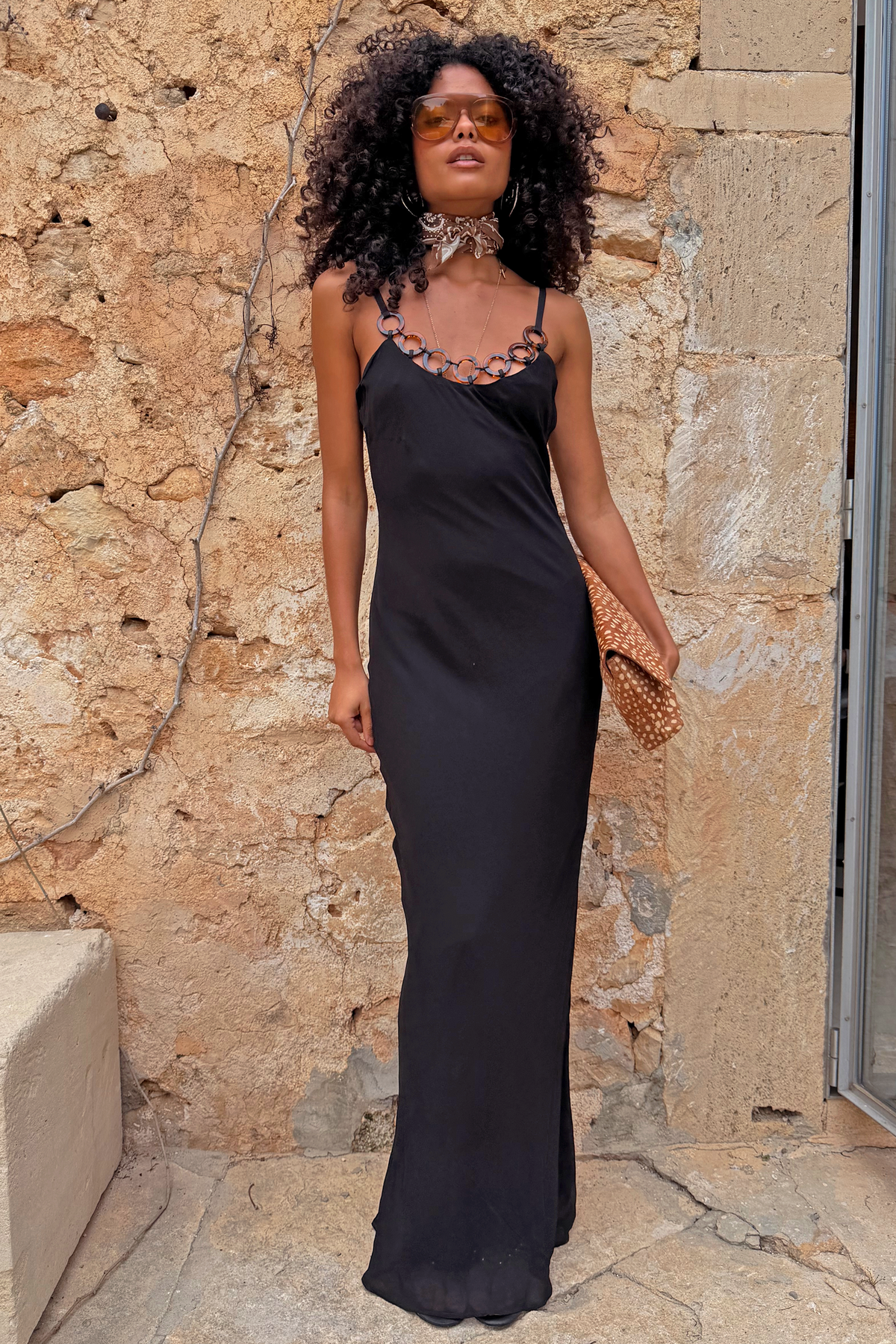 Black Sapna Petite maxi dress with ring detail neckline, styled with a spotted clutch against a stone wall.