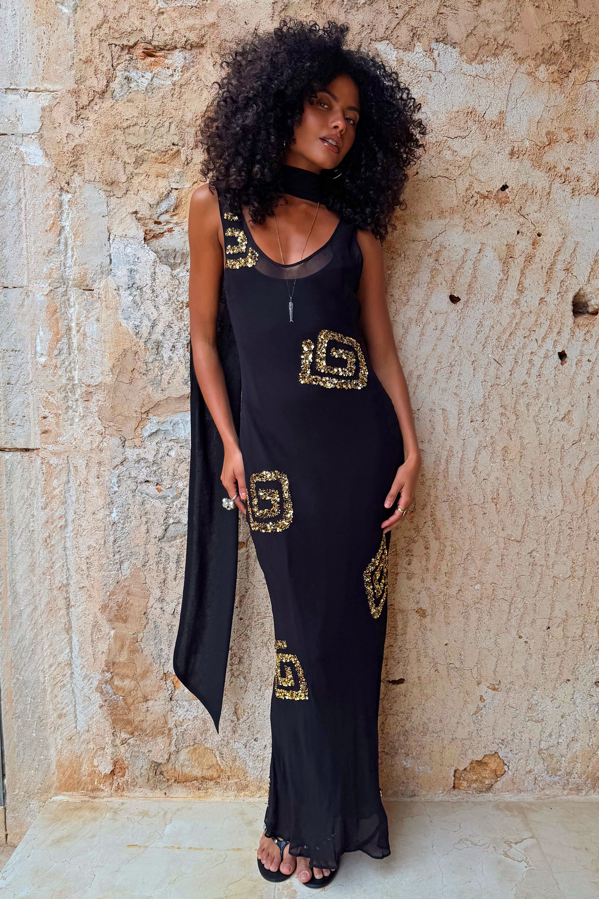 Black and gold Simona petite maxi dress with sequin patterns and a detachable scarf against a stone wall.