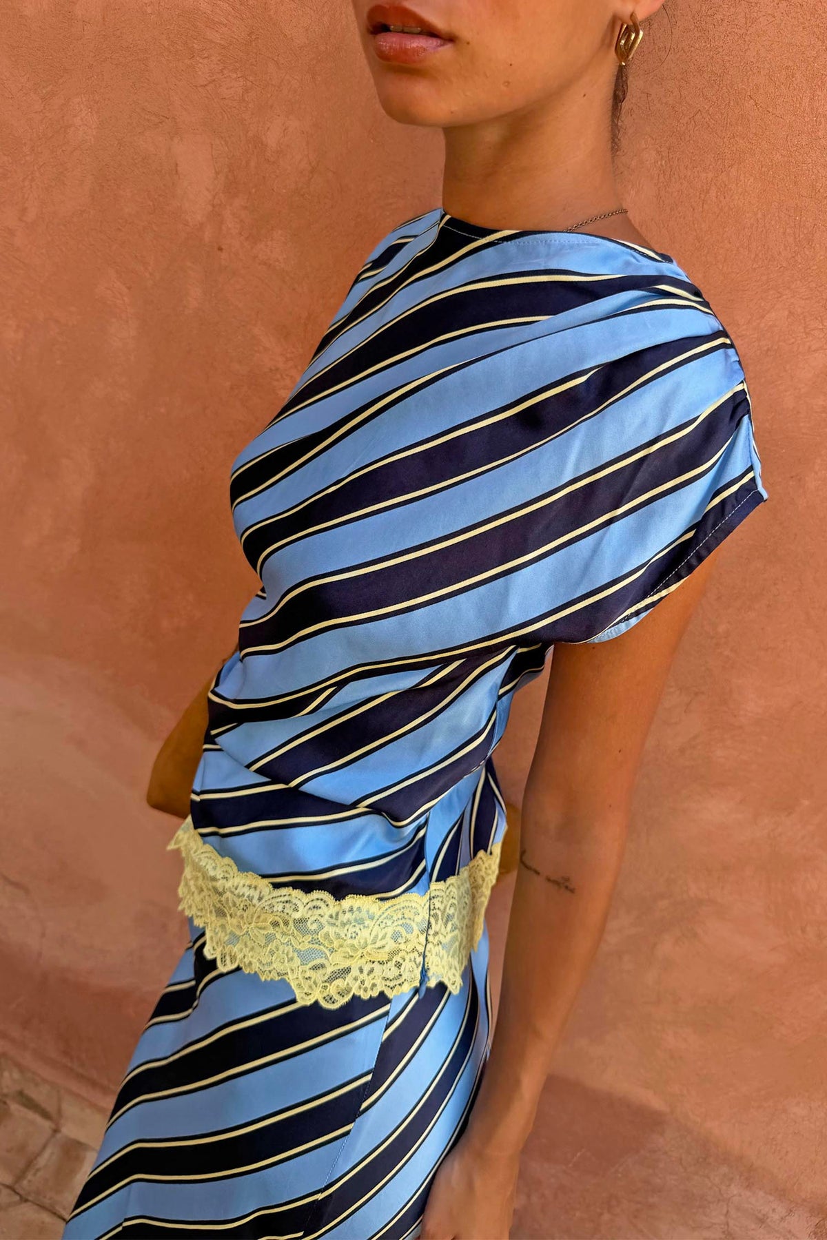 Stripe A-Symmetrical Top with blue diagonal stripes and a cream lace hem on a model against a peach wall.
