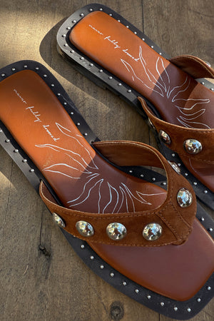 Tan leather stud sandals with silver studs, a square toe and suede straps on a wooden background.