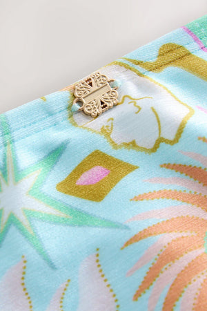Blue Dreamer High Waist Brief detail with a gold metal NFD logo and sun and palm motifs on blue swim fabric.