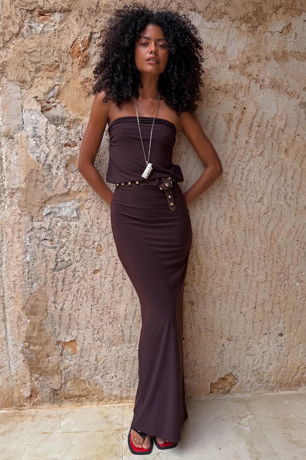 Chocolate Sabine Dress in brown, styled as a strapless column maxi dress on a model against a rustic wall.