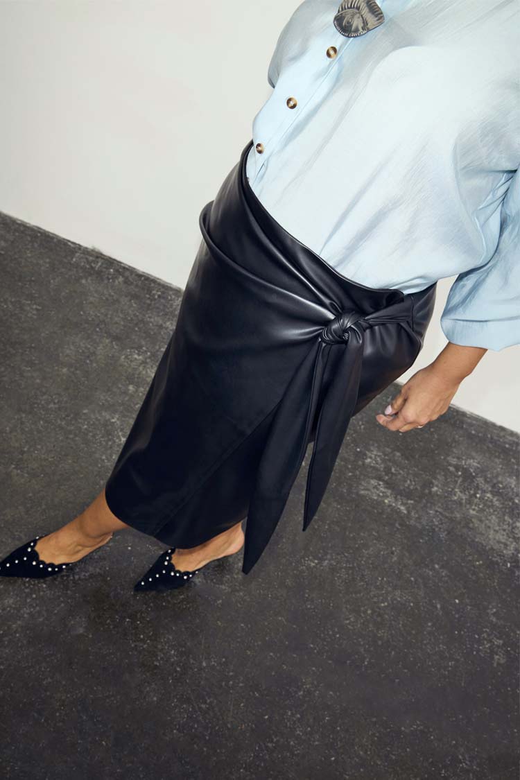 Iconic Black Vegan Leather Wrap Skirt with a tulip shaped tie, styled with a blue shirt and studded black mules.
