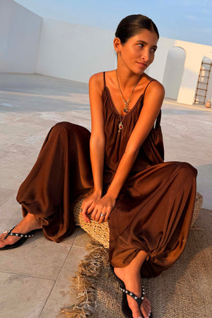 Chocolate Balloon Jumpsuit with a draped silhouette and gathered neckline, shown on a model sitting on a woven stool.