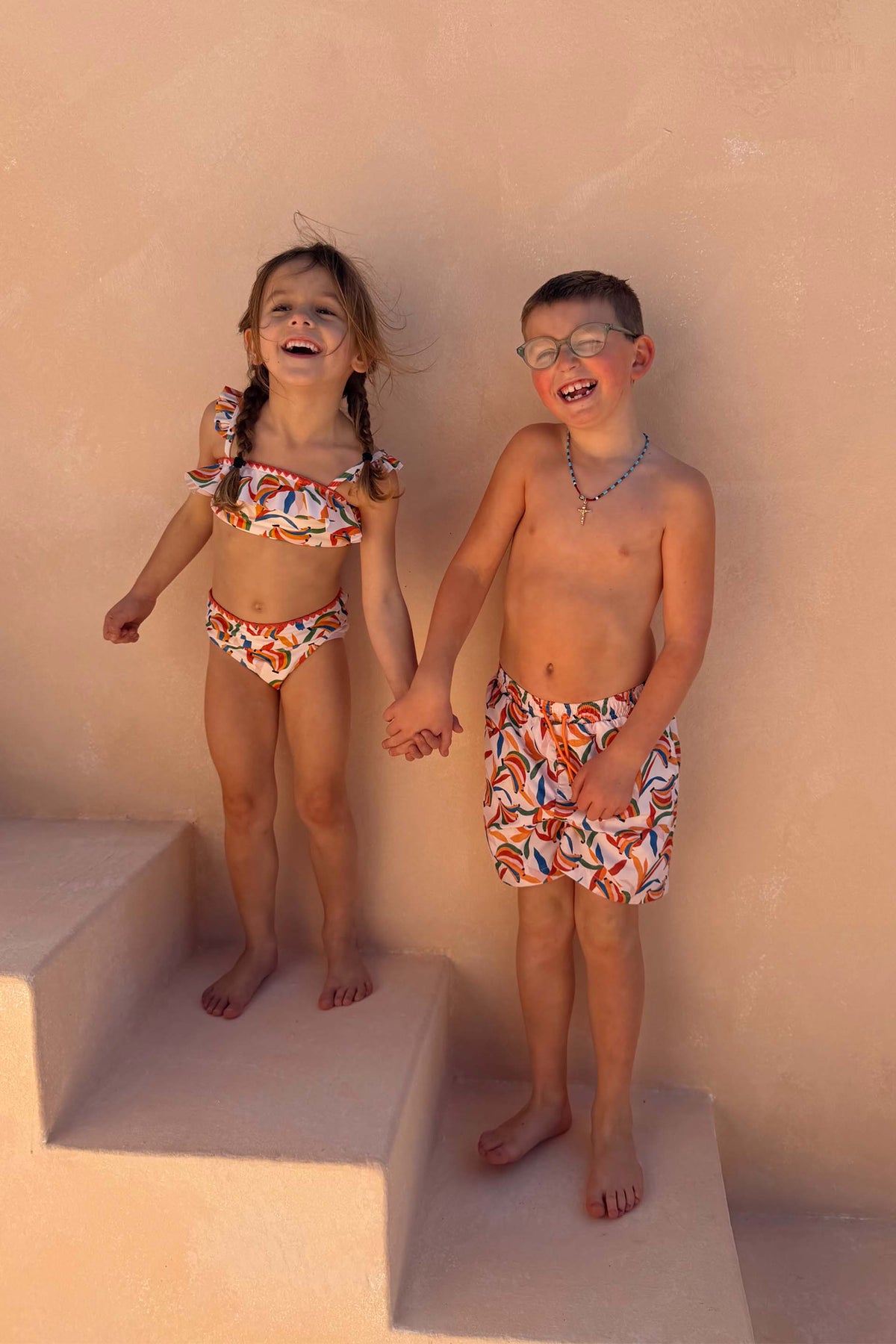 Boy's Banana Print Swim Shorts in cream with a multi-coloured print, worn by a child laughing on stairs.