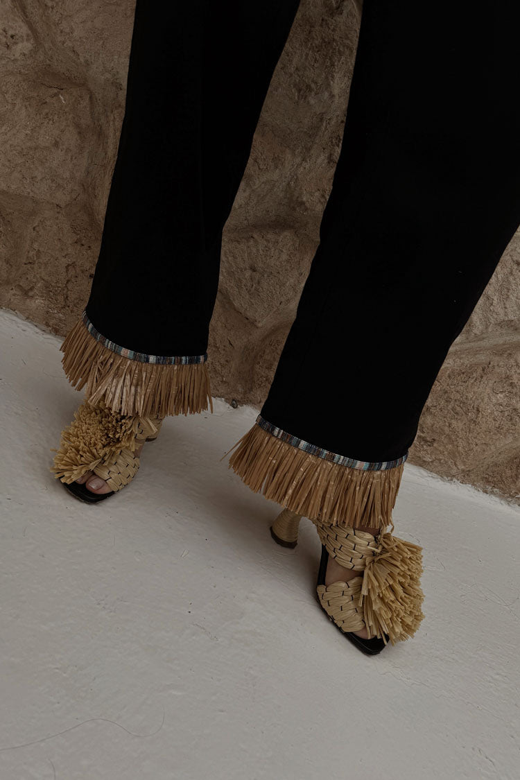 Black Raffia Marra Trousers featuring tan raffia fringe at the hem, styled with woven raffia sandals.