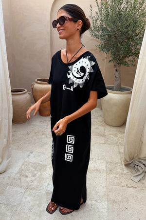Black Dolce Vita cotton maxi dress with white sun motifs and Greek graphics in an outdoor terrace setting.
