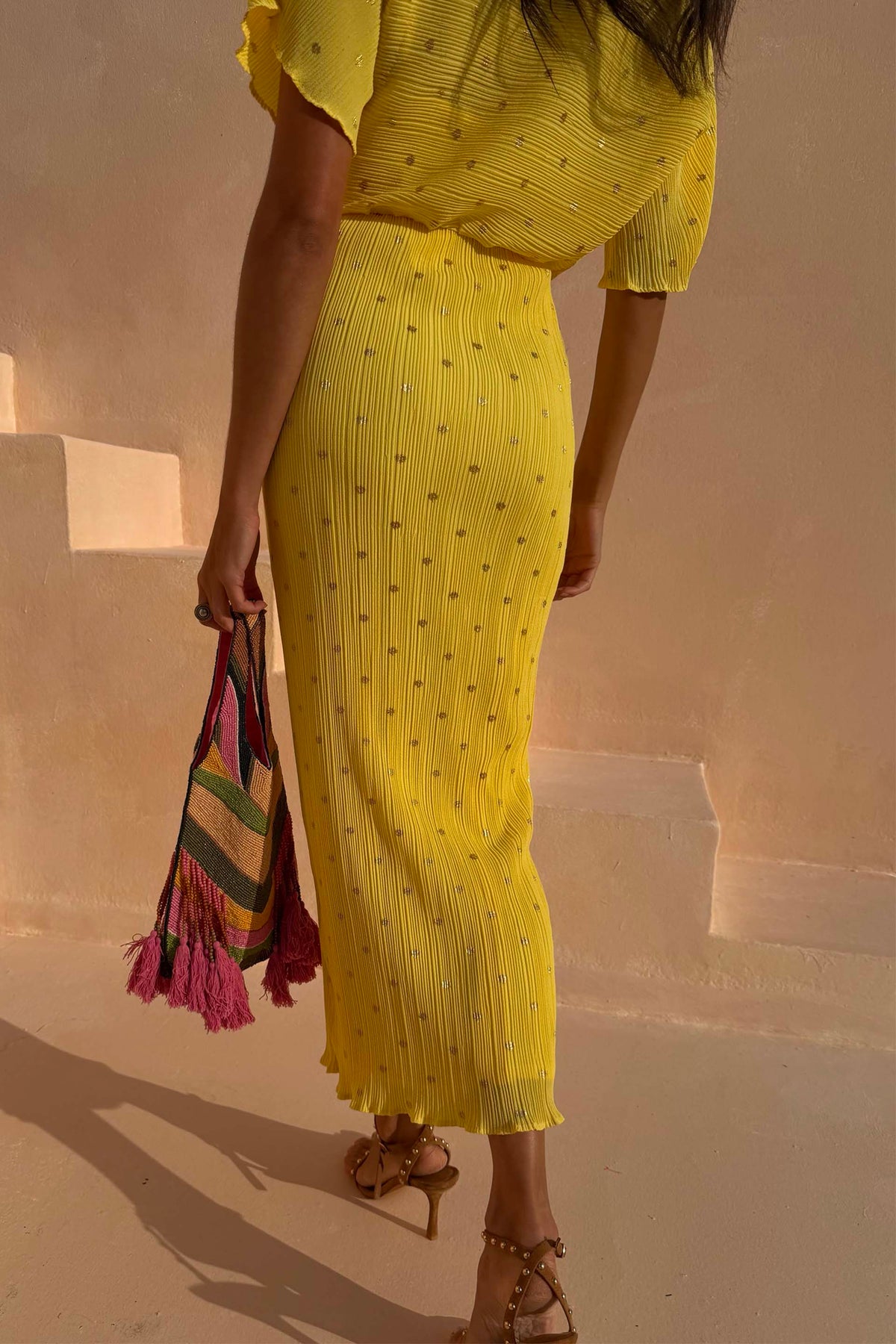 Sunshine Tilly Dress in yellow plissé with gold flecks and lettuce hem, held with a colourful beaded bag.