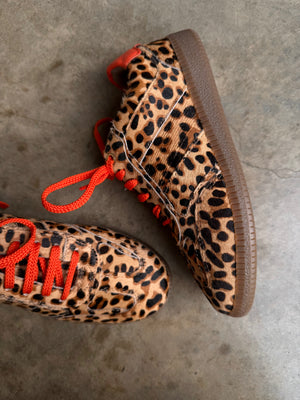 Leopard Pony Trainer featuring textured leopard print, vibrant orange laces, and a gum sole on a concrete surface.