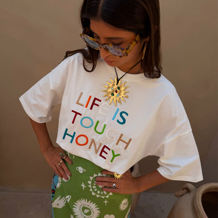 White Life Is Tough T-Shirt with colourful embroidered slogan, paired with a green patterned skirt and gold sun jewellery.