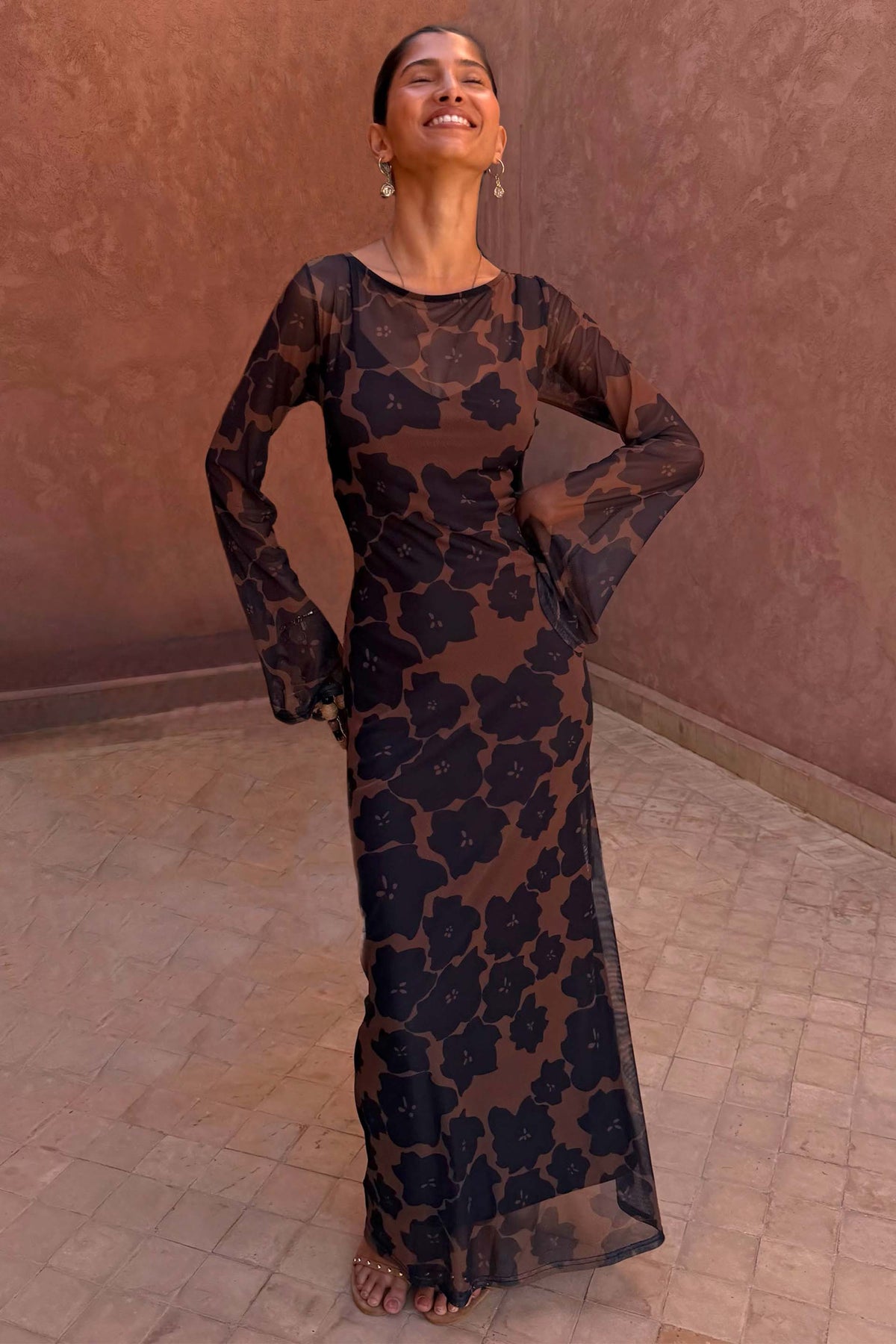 Tan Jaden Floral Mesh Maxi Dress with flared sleeves and a high neckline, worn against a terracotta background.