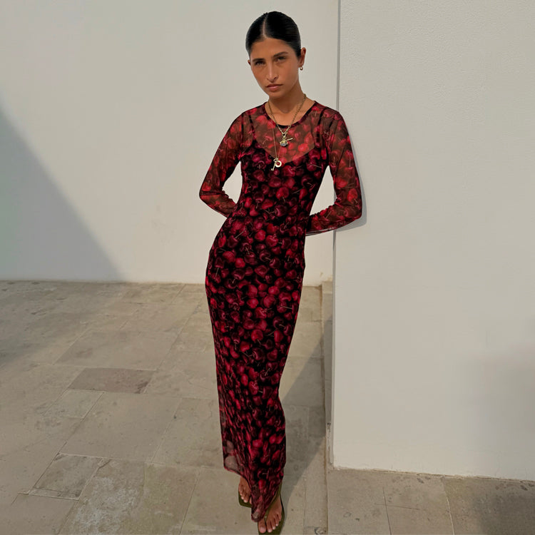 Red digital cherry print Mesh Soho Dress with long sleeves, designed in London and worn by a model against a wall.