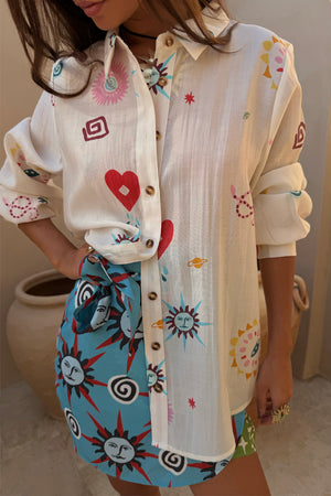 Sheer cream Morocco Charms Marley Shirt with heart and sun motifs, styled with a vibrant blue printed wrap skirt.