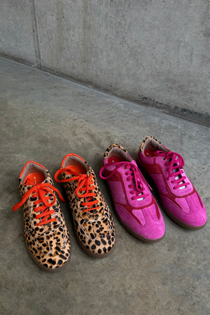Pink and red leather trainers beside leopard print sneakers with orange laces on a grey concrete background.