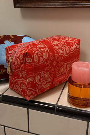 Red Bandana Reversible Make Up Bag with a cream paisley print and gold zipper, shown on a tiled bathroom ledge.