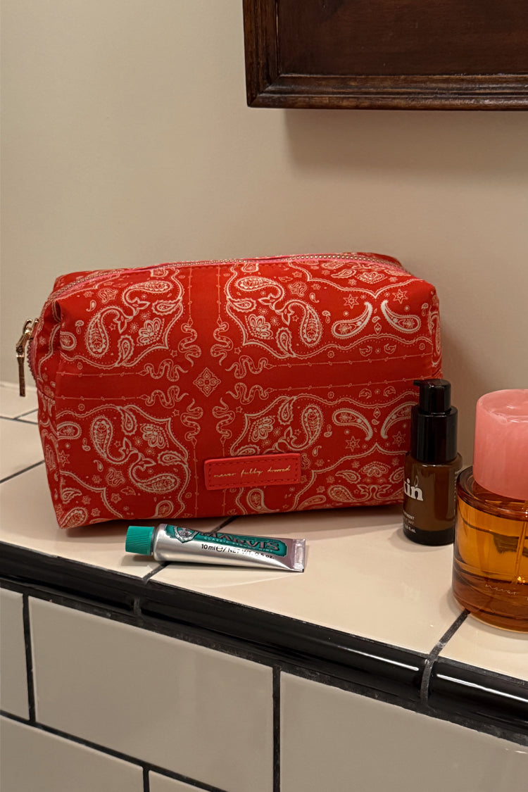 Red and pink bandana print makeup bag with a gold zip, shown on a tiled bathroom counter next to skincare essentials.