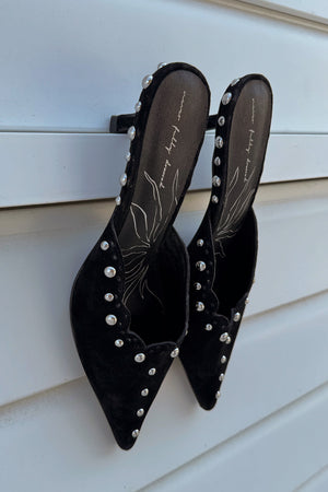 Black suede mules with silver stud detailing and scalloped edges, leaning against a white slatted background.
