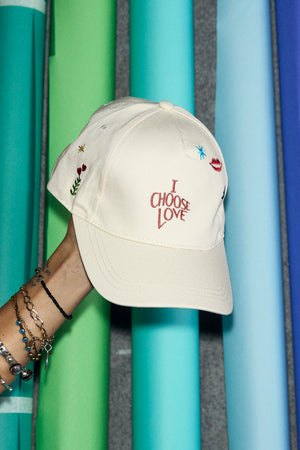 Cream Choose Love Cap with red 'I Choose Love' embroidery and playful motifs held against colourful paper rolls.