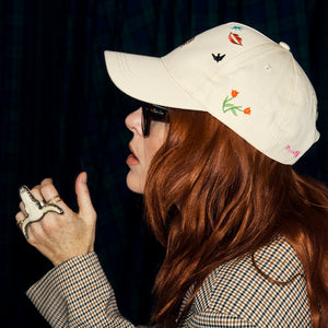Cream Choose Love Cap with colourful embroidered symbols, worn by a model with red hair.