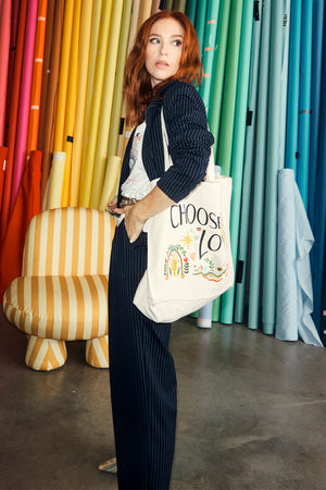 Choose Love Tote Bag with colourful graphics, held by Angela Scanlon in a navy pinstripe suit against a rainbow backdrop.