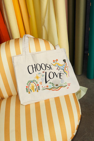 Choose Love Tote Bag in cream recycled cotton with colourful graphic prints, resting on a yellow striped armchair.