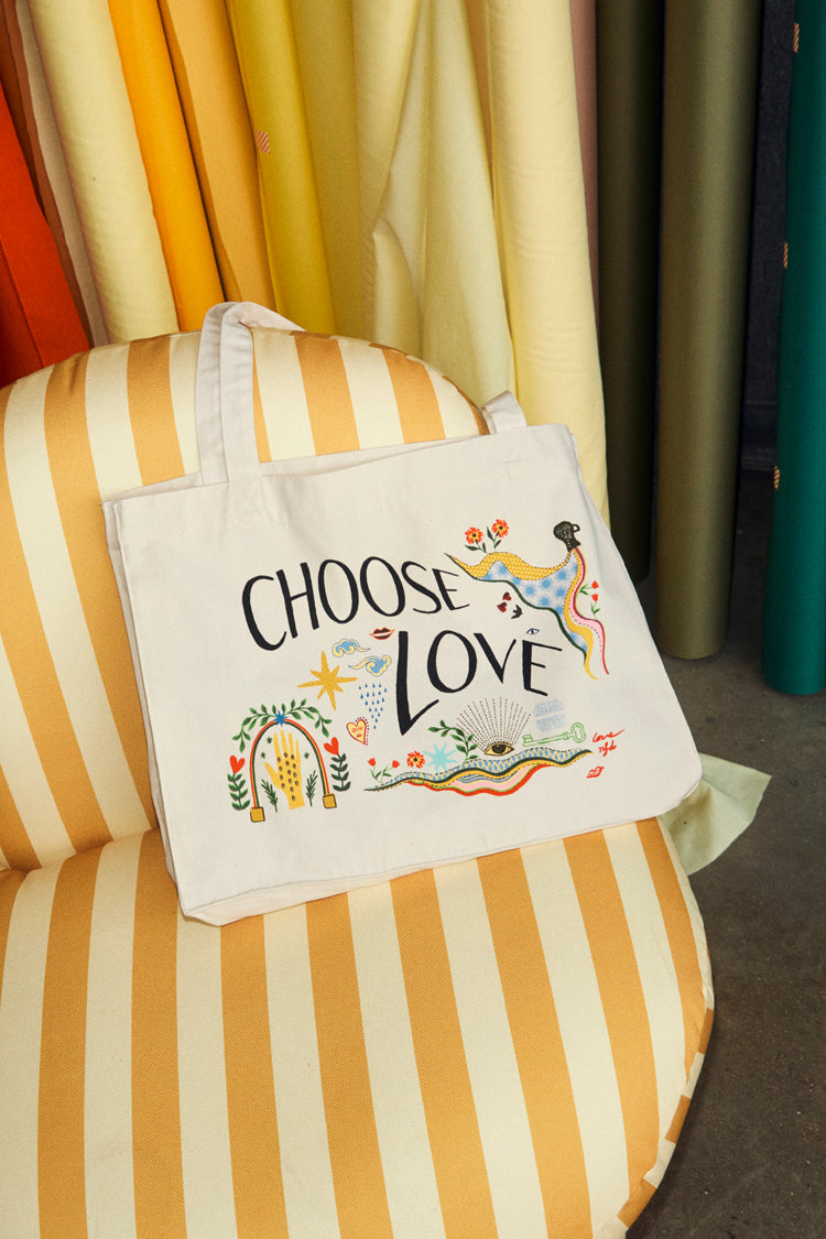Choose Love Tote Bag in cream recycled cotton with colourful graphic prints, resting on a yellow striped armchair.