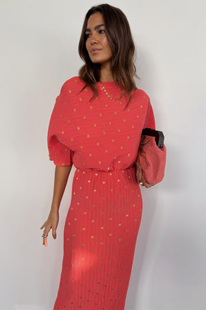 Coral Plisse Tilly Dress featuring gold flecks, an off-the-shoulder neckline and elasticated waist, styled with a pink clutch.