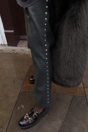 Bottle green vegan leather trousers with silver side studs, styled with black buckled shoes on a stone pavement.