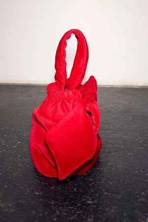 Red Velvet Bow Bag with an oversized bow detail and padded top handle, set against a dark textured floor.