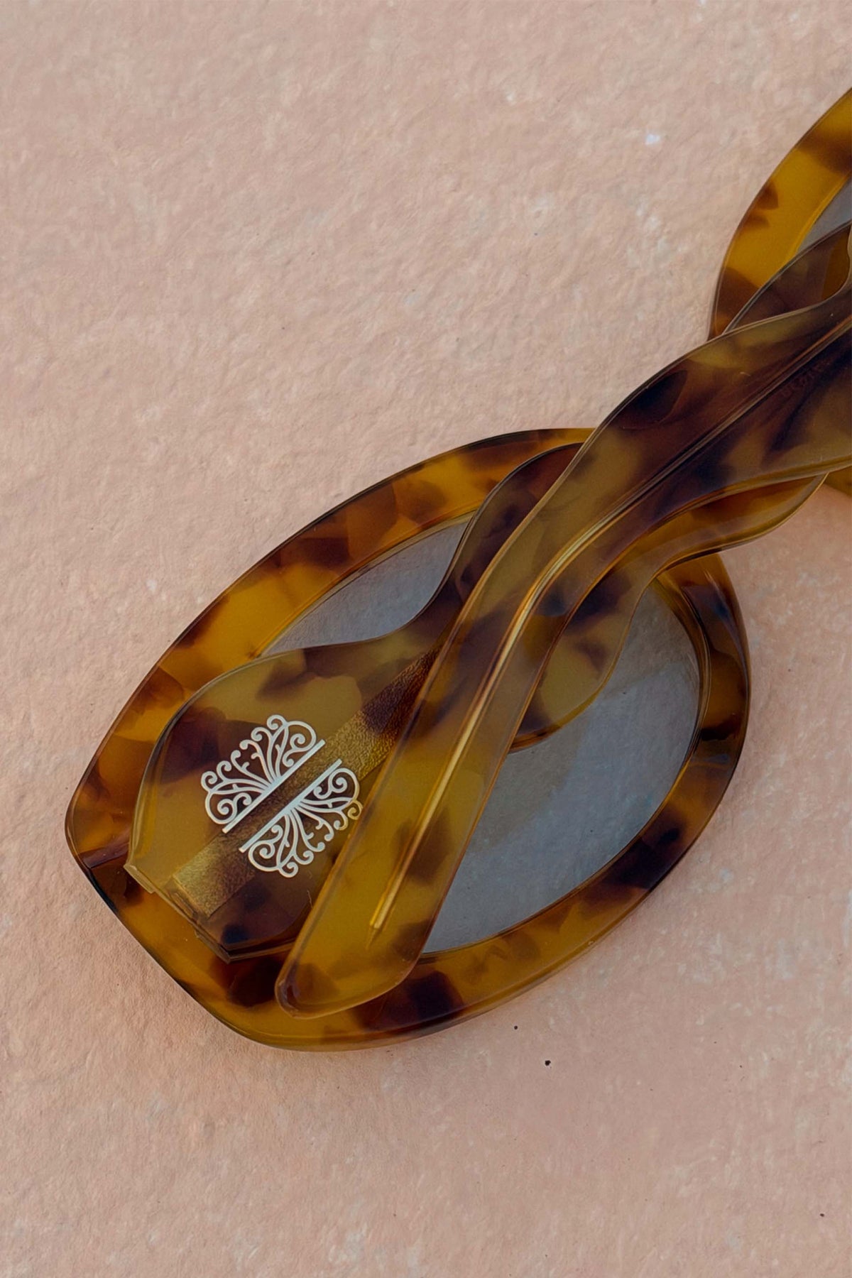 Occhiali da sole tartarugati gialli con aste a onda e dettaglio logo su sfondo naturale.