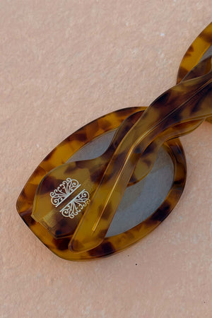Occhiali da sole tartarugati gialli con aste a onda e dettaglio logo su sfondo naturale.